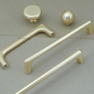 May include: A collection of gold-colored cabinet hardware, including two knobs and three different handle styles. The hardware has a smooth, matte finish and is designed for furniture or cabinetry.