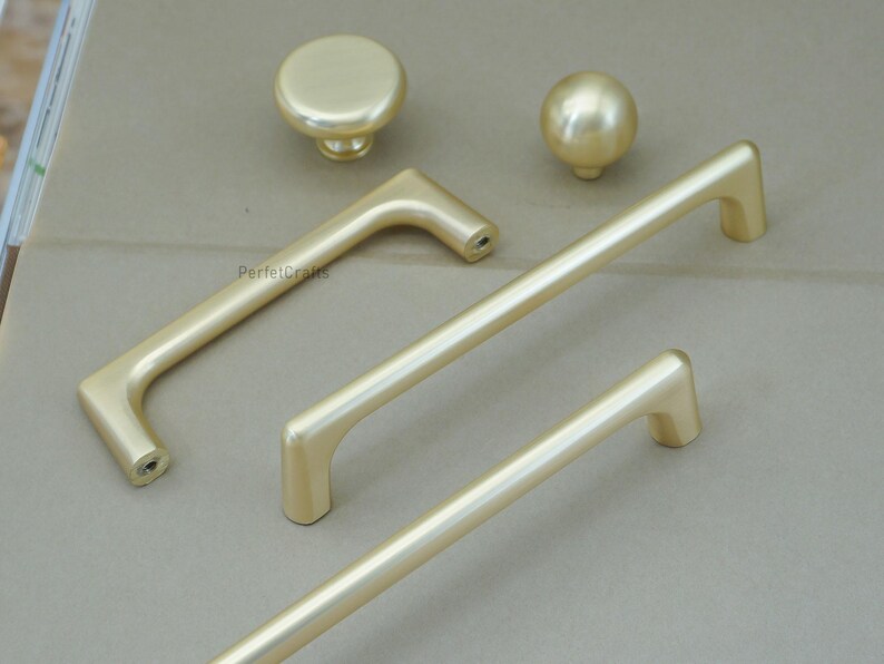 May include: A collection of gold-colored cabinet hardware, including two knobs and three different handle styles. The hardware has a smooth, matte finish and is designed for furniture or cabinetry.