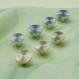May include: A collection of silver and gold-toned cabinet knobs with a radial design on the circular top. The knobs are arranged on a light green fabric. The brand name "Perfetcrafts" is printed on the fabric.