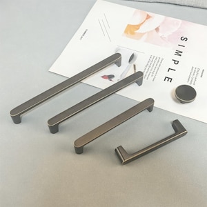 May include: A selection of bronze-coloured cabinet handles, comprising rectangular bar pulls of different lengths and a round knob. The hardware is arranged on a light grey surface, with a magazine page visible in the background.