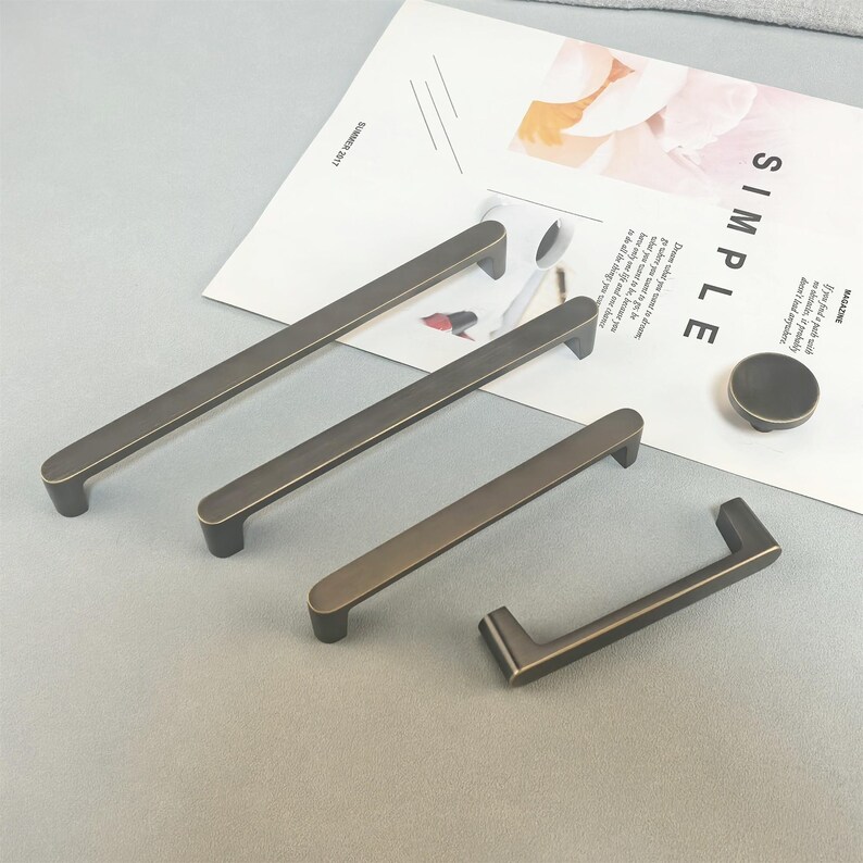 May include: A selection of bronze-coloured cabinet handles, comprising rectangular bar pulls of different lengths and a round knob. The hardware is arranged on a light grey surface, with a magazine page visible in the background.