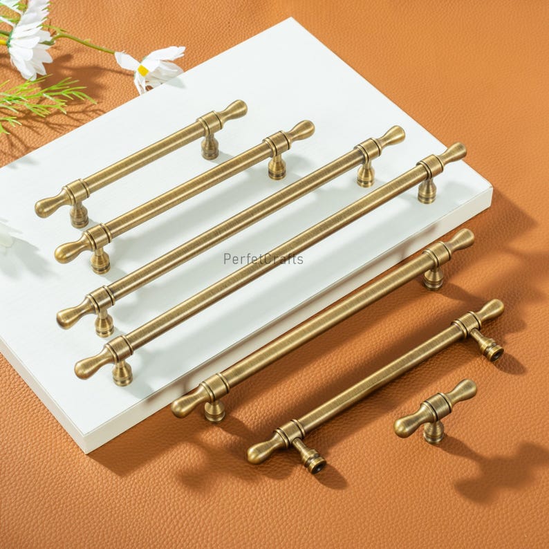 May include: A selection of antique brass cabinet pulls and handles are displayed. The hardware has a T-bar design with a vintage appearance. The pulls vary in length, offering a range of sizes for various cabinet applications. The longest pull is approximately 20 cm.