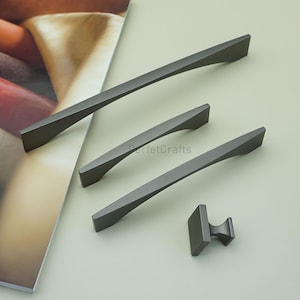 May include: A set of four dark gray cabinet pulls and a knob. The pulls are in varying lengths, with a curved, angular design. The knob is square with a curved handle. The items are arranged on a light green surface.