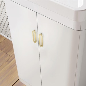 May include: A white cabinet with two gold oval handles. The handles are vertically aligned and have a detailed design. The cabinet has a glossy finish. The cabinet is set against a light-colored wooden floor.