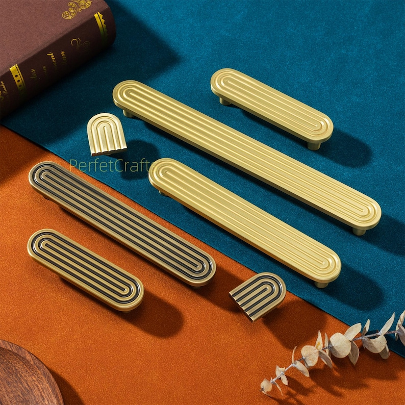 May include: A collection of gold and black cabinet hardware, including handles and decorative pieces. The handles have a grooved design and are in various sizes. The hardware is displayed on a blue and orange surface.