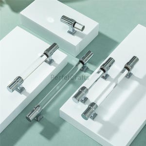 May include: A collection of cabinet hardware featuring clear acrylic handles with polished chrome accents. The handles are rectangular and cylindrical, with a modern design. The hardware is displayed on white platforms against a light green background.