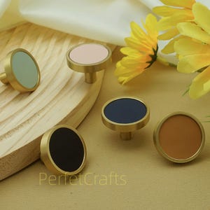 Puede incluir: Un conjunto de cinco pomos de armario con bases de color lat&oacute;n y tapas circulares en varios colores: negro, azul marino, rosa claro, verde salvia y marr&oacute;n. Los pomos est&aacute;n dispuestos sobre una superficie de madera, con flores amarillas en el fondo.