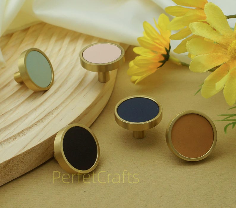 Puede incluir: Un conjunto de cinco pomos de armario con bases de color lat&oacute;n y tapas circulares en varios colores: negro, azul marino, rosa claro, verde salvia y marr&oacute;n. Los pomos est&aacute;n dispuestos sobre una superficie de madera, con flores amarillas en el fondo.