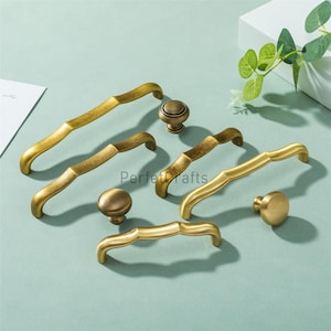 May include: An assortment of antique brass cabinet hardware, including handles and knobs. The handles have a curved design with a bamboo-like texture. The knobs are round and solid. The hardware is arranged on a light blue surface.