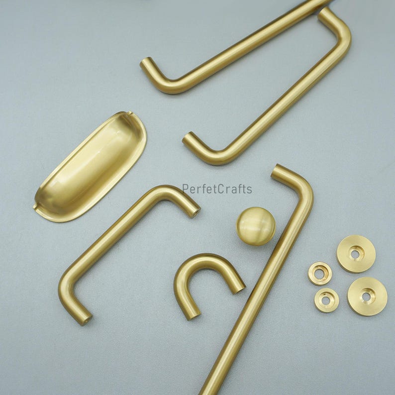 May include: An assortment of gold-coloured cabinet hardware, including handles, knobs, and pulls. The hardware is displayed on a grey surface, showcasing various shapes and sizes. The brand name "PerfetcCrafts" is visible on the image.