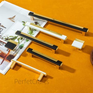May include: A collection of modern cabinet hardware, including black and gold, white and gold, and black and white handles and knobs. The hardware is displayed on a white surface with the text "PerfetcCrafts" in the bottom center.