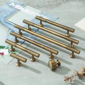 May include: A selection of antique brass cabinet handles and a knob. The handles are long bar pulls with a ribbed design, and the knob has a similar pattern. The hardware is displayed on a light surface.