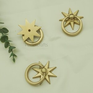May include: Three gold-coloured starburst drawer pulls with ring handles. The pulls are arranged on a light-coloured surface. The text "PerfectCrafts" is visible on one of the pulls.