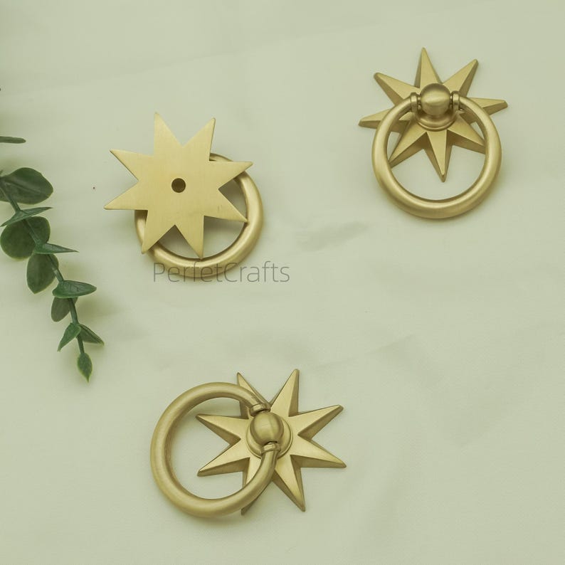 May include: Three gold-coloured starburst drawer pulls with ring handles. The pulls are arranged on a light-coloured surface. The text "PerfectCrafts" is visible on one of the pulls.