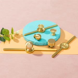 Puede incluir: Una colecci&oacute;n de herrajes dorados para armarios, que incluyen pomos y tiradores. Los herrajes presentan varios dise&ntilde;os, como pomos esf&eacute;ricos, cil&iacute;ndricos y en forma de hongo, y tiradores de barra. El texto "PerfecCrafts" es visible.