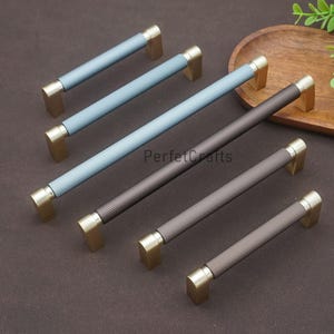 Puede incluir: Un conjunto de tiradores de armario en varios tamaños y colores. Los tiradores tienen una textura acanalada en tonos azules y marrones, con extremos rectangulares dorados. El texto "PerfectCrafts" es visible.