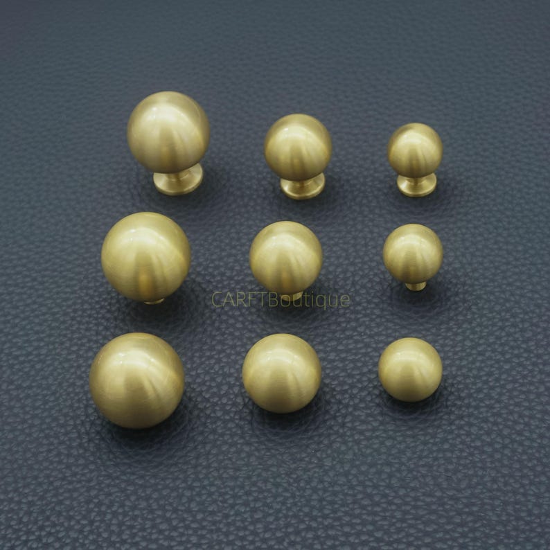 Puede incluir: Nueve pomos de lat&oacute;n para armarios de diferentes tama&ntilde;os est&aacute;n dispuestos sobre una superficie oscura y texturizada. Cada pomo es esf&eacute;rico con una peque&ntilde;a base redonda. El lat&oacute;n tiene un acabado mate, que refleja la luz.