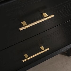 K&ouml;nnte beinhalten: Nahaufnahme einer schwarzen Holzkommode mit zwei Schubladen. Jede Schublade hat einen goldfarbenen, zylindrischen Griff mit rechteckigen Montageplatten. Die Griffe sind horizontal ausgerichtet.