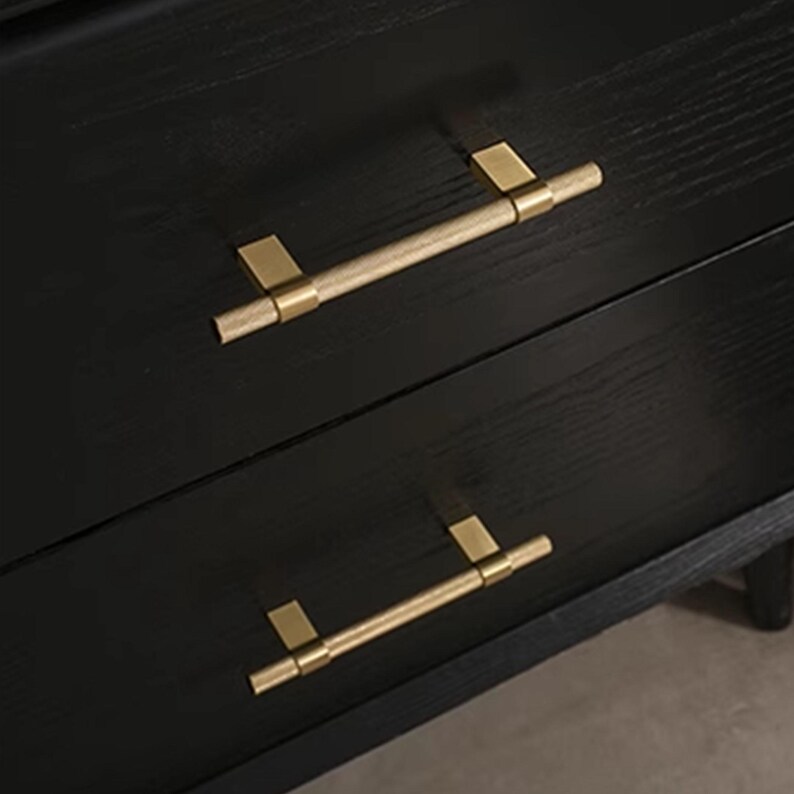 K&ouml;nnte beinhalten: Nahaufnahme einer schwarzen Holzkommode mit zwei Schubladen. Jede Schublade hat einen goldfarbenen, zylindrischen Griff mit rechteckigen Montageplatten. Die Griffe sind horizontal ausgerichtet.