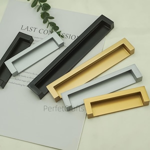 Peut inclure: Plusieurs poignées de meubles rectangulaires de couleurs variées, dont noir, argent et or. Les poignées sont en métal et présentent un design encastré. Le texte "LAST COMSSIO" est visible sur un papier en arrière-plan.