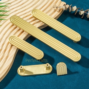 May include: A collection of gold-colored cabinet hardware, including handles and a small decorative piece. The handles have a rectangular shape with a grooved design. The hardware is arranged on a wooden surface and a blue background.