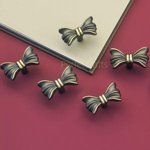 Może przedstawiać: Sześć dekoracyjnych gałek do szafek w kształcie kokardki, z ciemnobrązowym wykończeniem i złotymi akcentami. Gałki są ułożone na czerwonej powierzchni z beżowo-czarnym prostokątnym przedmiotem w tle. Widoczny jest tekst "Perfect Crafts".