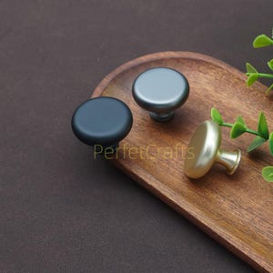 以下が含まれることがあります： マットブラック、ガンメタルグレー、つや消しゴールドの3種類の仕上げのキャビネットノブ。ノブは丸く、シンプルでモダンなデザインです。「PerfecCrafts」の文字が木製のトレイに表示されています。