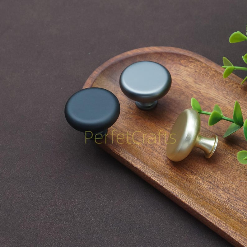 以下が含まれることがあります： マットブラック、ガンメタルグレー、つや消しゴールドの3種類の仕上げのキャビネットノブ。ノブは丸く、シンプルでモダンなデザインです。「PerfecCrafts」の文字が木製のトレイに表示されています。