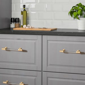 May include: Close-up of gray kitchen cabinet drawers with gold and clear acrylic bar pulls. The drawers are part of a kitchen setup with a black countertop, white subway tile backsplash, and a small potted plant.