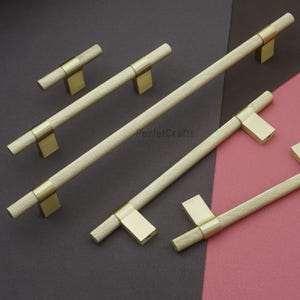 K&ouml;nnte beinhalten: Eine Sammlung goldfarbener Schrankgriffe und -kn&ouml;pfe. Die Griffe haben einen strukturierten Griff mit Rautenmuster und sind auf rechteckigen Sockeln montiert. Die Kn&ouml;pfe haben ein &auml;hnliches Design. Die Hardware wird vor einem dunkelgrauen und rosa Hintergrund pr&auml;sentiert.