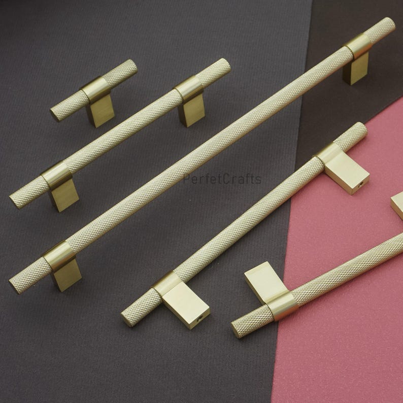 K&ouml;nnte beinhalten: Eine Sammlung goldfarbener Schrankgriffe und -kn&ouml;pfe. Die Griffe haben einen strukturierten Griff mit Rautenmuster und sind auf rechteckigen Sockeln montiert. Die Kn&ouml;pfe haben ein &auml;hnliches Design. Die Hardware wird vor einem dunkelgrauen und rosa Hintergrund pr&auml;sentiert.