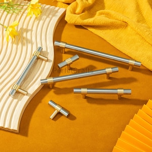 Puede incluir: Un conjunto de tiradores de armario con un diseño moderno. Los tiradores presentan una combinación de acabados plateados y dorados cepillados. Los tiradores cilíndricos están acentuados con bandas doradas, y los postes de montaje también son dorados. Sobre fondo amarillo.