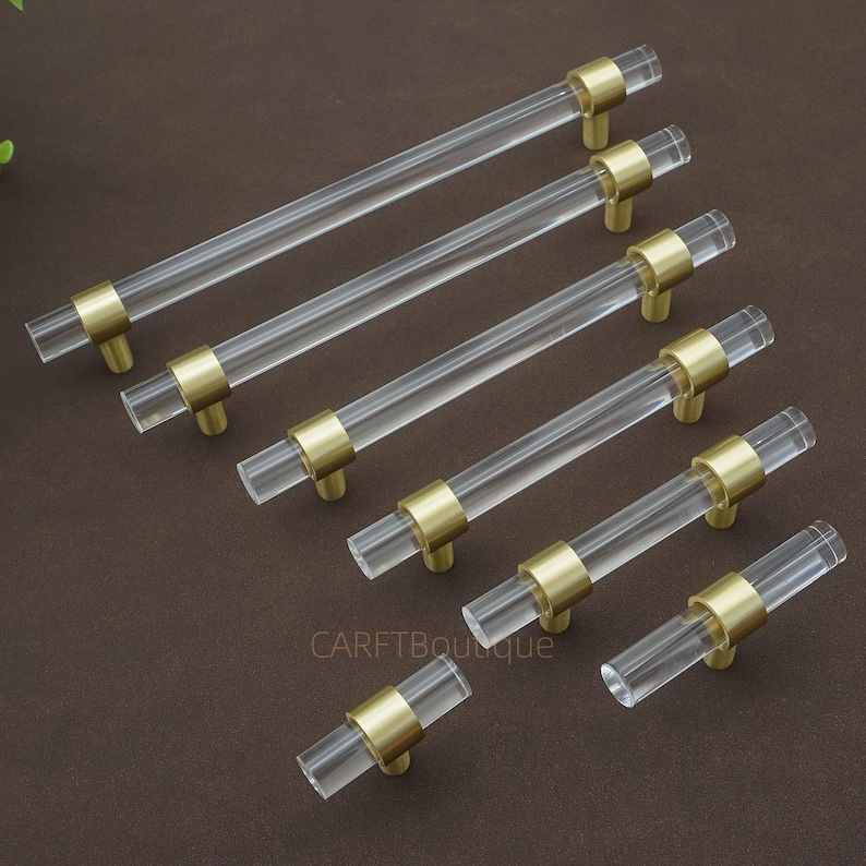 May include: Clear acrylic cabinet pulls with brass-coloured accents. The pulls are cylindrical and have a modern design. The hardware is arranged on a brown surface. The text "CARFTBoutique" is visible on the surface.