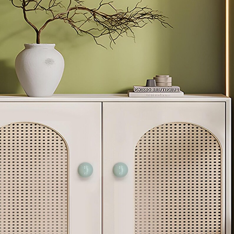 May include: A cream-coloured cabinet with two doors, each with a woven rattan panel in an arched design. The cabinet has two pale blue round knobs. A white vase with a branch sits on top, along with books and ornaments.