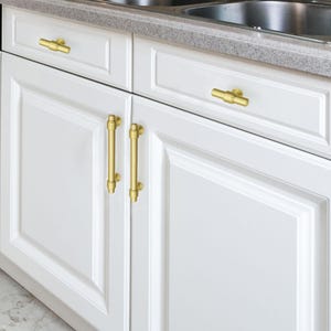 Peut inclure: Des armoires de cuisine blanches avec des ferrures dor&eacute;es. Les armoires ont une conception &agrave; panneaux encastr&eacute;s et sont accentu&eacute;es de barres et de poign&eacute;es dor&eacute;es. Le comptoir est de couleur gris clair.