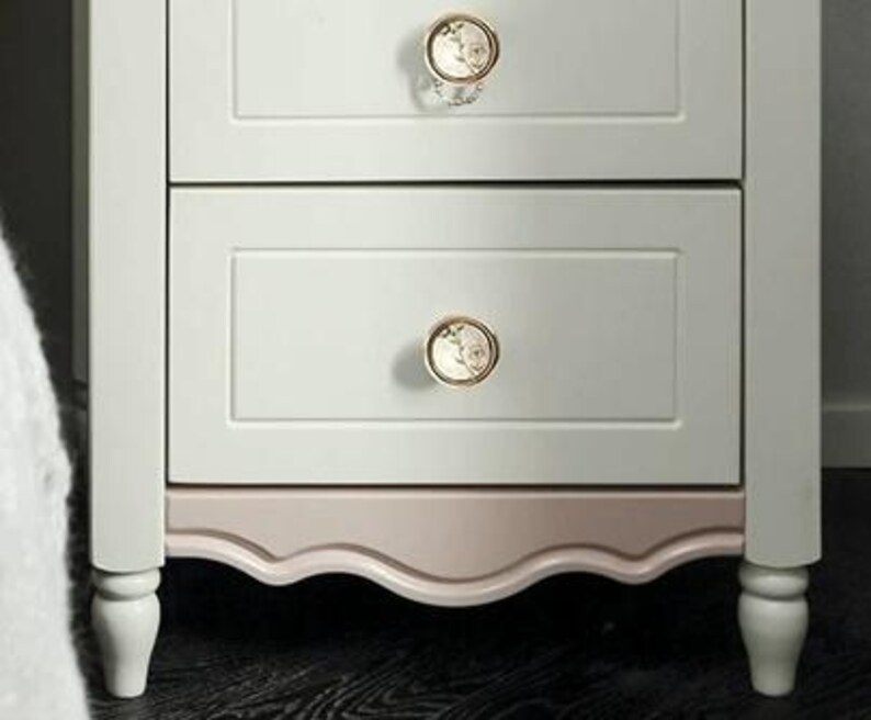 Pu&ograve; includere: Un comodino a due cassetti dipinto in bianco sporco e rosa chiaro. I cassetti hanno pomelli decorativi floreali con finiture dorate. Il bordo inferiore del comodino ha un design smerlato.