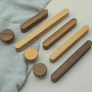 May include: A collection of wooden cabinet knobs and pulls in various shades of brown. The knobs are round, while the pulls are rectangular with rounded ends. The wood grain is visible, and the items are arranged on a light gray surface.
