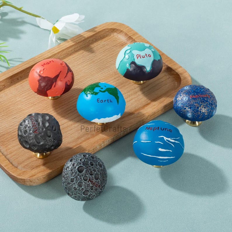May include: A collection of decorative cabinet knobs, each designed to resemble a planet in our solar system. The knobs feature detailed paintwork, including the names of the planets. The knobs are mounted on brass-colored bases.