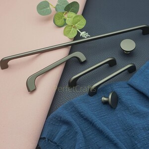 Op de afbeelding: Een verzameling donkergrijze kastbeslagen, waaronder handgrepen en knoppen in verschillende maten. De handgrepen hebben een gebogen ontwerp en de knoppen zijn rond. De hardware wordt gepresenteerd op een roze en blauwe stoffen achtergrond.