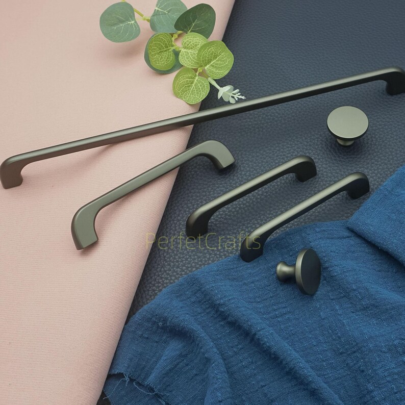 Op de afbeelding: Een verzameling donkergrijze kastbeslagen, waaronder handgrepen en knoppen in verschillende maten. De handgrepen hebben een gebogen ontwerp en de knoppen zijn rond. De hardware wordt gepresenteerd op een roze en blauwe stoffen achtergrond.