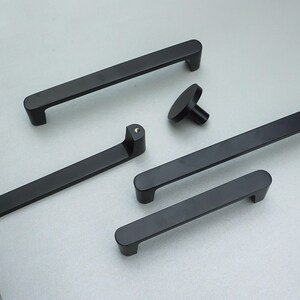May include: A selection of matte black cabinet handles and a knob. The handles are of varying lengths, with rounded ends and a modern design. The hardware is displayed on a light grey surface. The longest handle is approximately 30 cm.