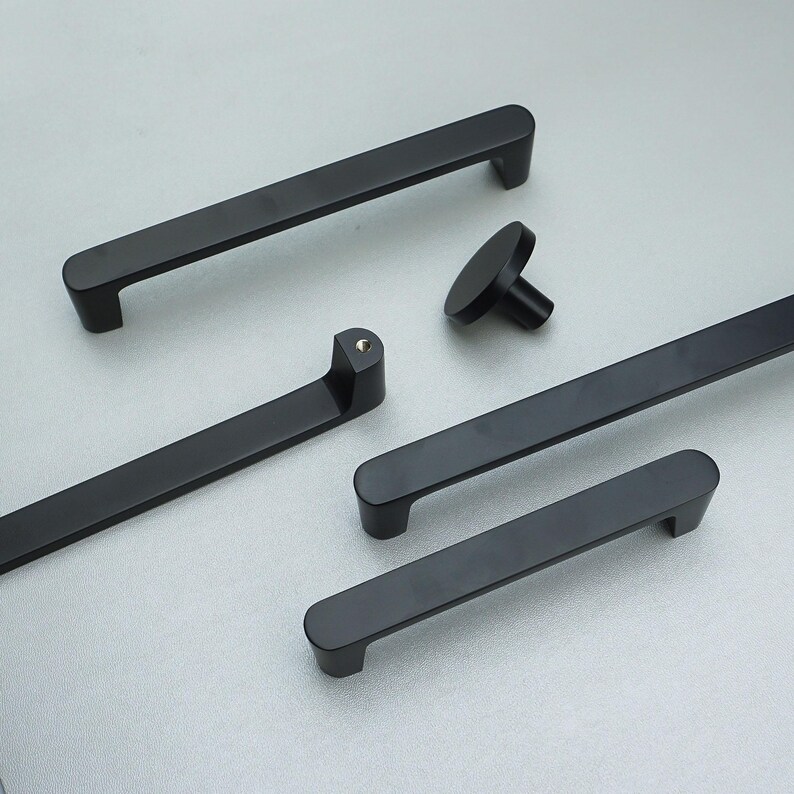 May include: A selection of matte black cabinet handles and a knob. The handles are of varying lengths, with rounded ends and a modern design. The hardware is displayed on a light grey surface. The longest handle is approximately 30 cm.