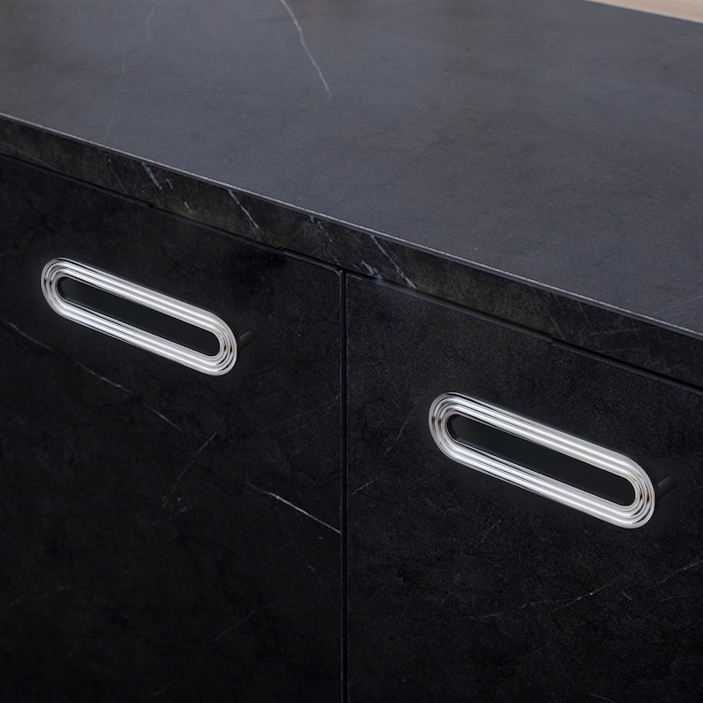 May include: A close-up of a black cabinet featuring two oval-shaped handles with silver rims. The cabinet has a dark, textured surface with white veining. The handles are positioned on the cabinet doors.