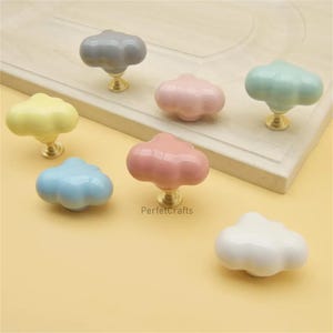 May include: A collection of cloud-shaped cabinet knobs in various pastel colors, including yellow, blue, pink, and green. Each knob has a gold-colored base. The knobs are displayed on a light-colored wooden surface.