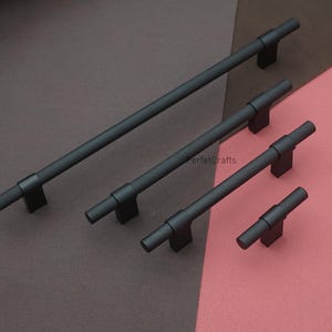 K&ouml;nnte beinhalten: Vier schwarze Schrankgriffe in verschiedenen Gr&ouml;&szlig;en. Jeder Griff hat einen strukturierten, zylindrischen Griff und eine rechteckige Basis. Sie sind auf einer braunen und rosa Oberfl&auml;che angeordnet.