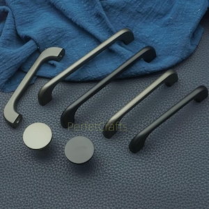 Puede incluir: Una selección de herrajes para armarios, que incluye tiradores y pomos redondos, en varios tamaños y acabados. Los herrajes son en tonos grises y negros, con un acabado mate. El texto "Perfectcrafts" es visible.