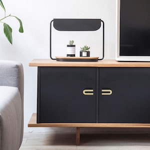 May include: A black cabinet with gold-coloured handles and a light wood top. A black metal frame with a small wooden tray holding two small potted plants sits on top. The cabinet is in a room with a white wall and a grey sofa.
