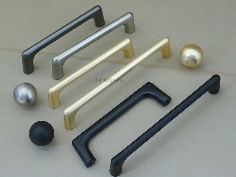 May include: A collection of cabinet hardware, including several bar pulls and round knobs. The pulls are in various finishes: dark gray, silver, gold, and black. The knobs are silver, gold, and black. The hardware is displayed on a neutral surface.