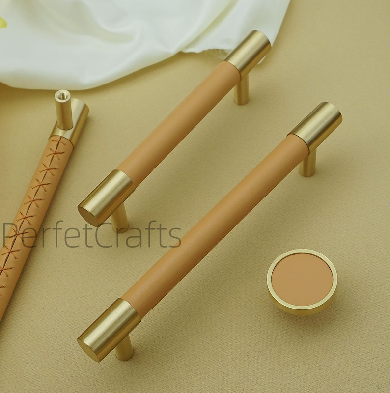 Puede incluir: Un conjunto de herrajes para armarios con tiradores envueltos en cuero marr&oacute;n y detalles dorados. Los tiradores son cil&iacute;ndricos y de dise&ntilde;o moderno. Tambi&eacute;n hay un pomo de armario redondo con una combinaci&oacute;n de colores a juego. El texto "PerfetcCrafts" es visible.