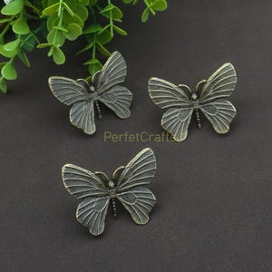 K&ouml;nnte beinhalten: Drei bronzefarbene Schrankgriffe in Schmetterlingsform. Jeder Schmetterling hat detaillierte Fl&uuml;gelmuster und einen strukturierten K&ouml;rper. Die Griffe sind auf einer dunklen Oberfl&auml;che angeordnet, mit Gr&uuml;n im Hintergrund. Der Text "PerfecCrafts" ist sichtbar.